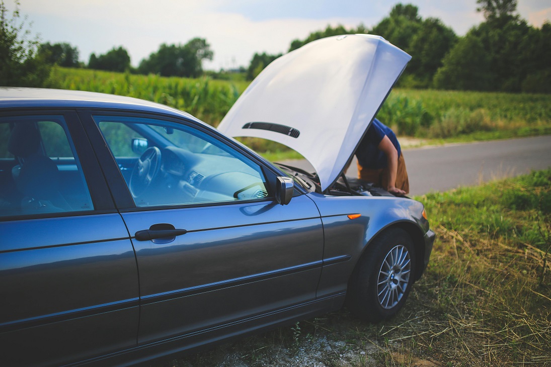 Persona revisando el motor de un automóvil con el capó abierto al costado de una carretera rural.