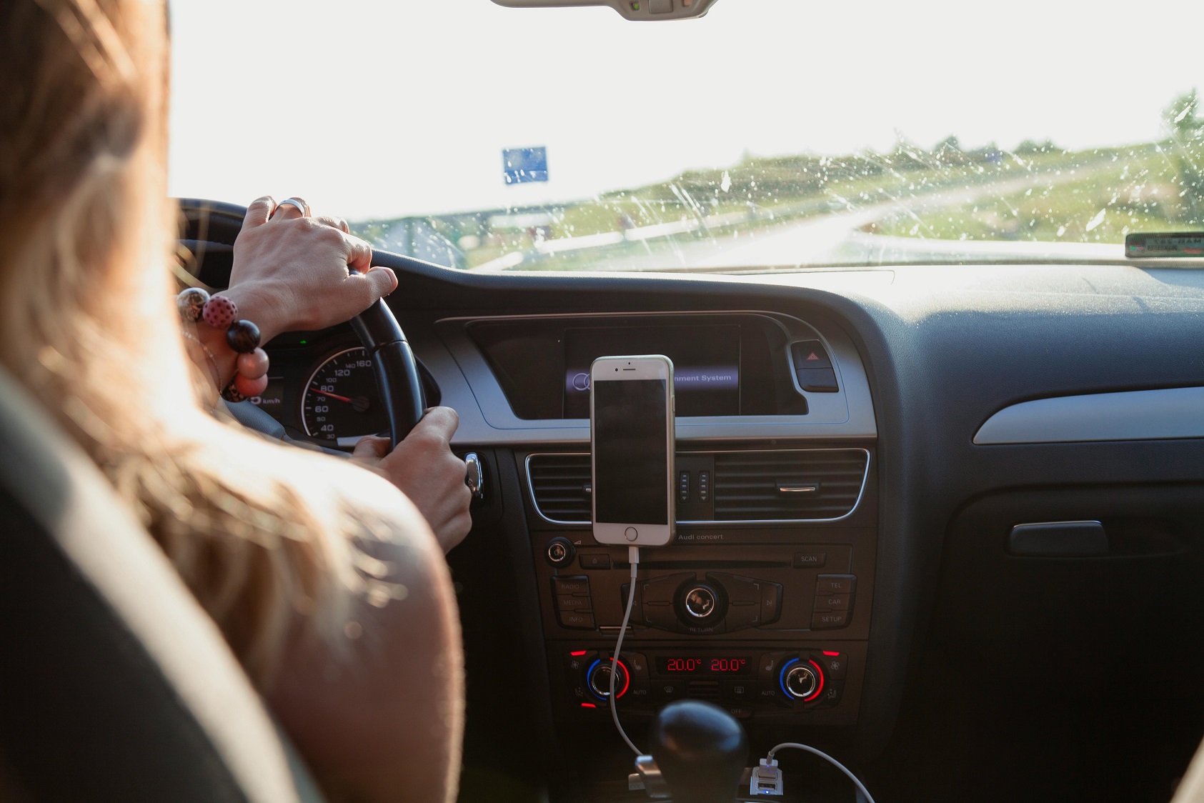 Mujer conduciendo un automóvil en una carretera soleada, con un teléfono móvil sujeto en el tablero y el camino visible al fondo.